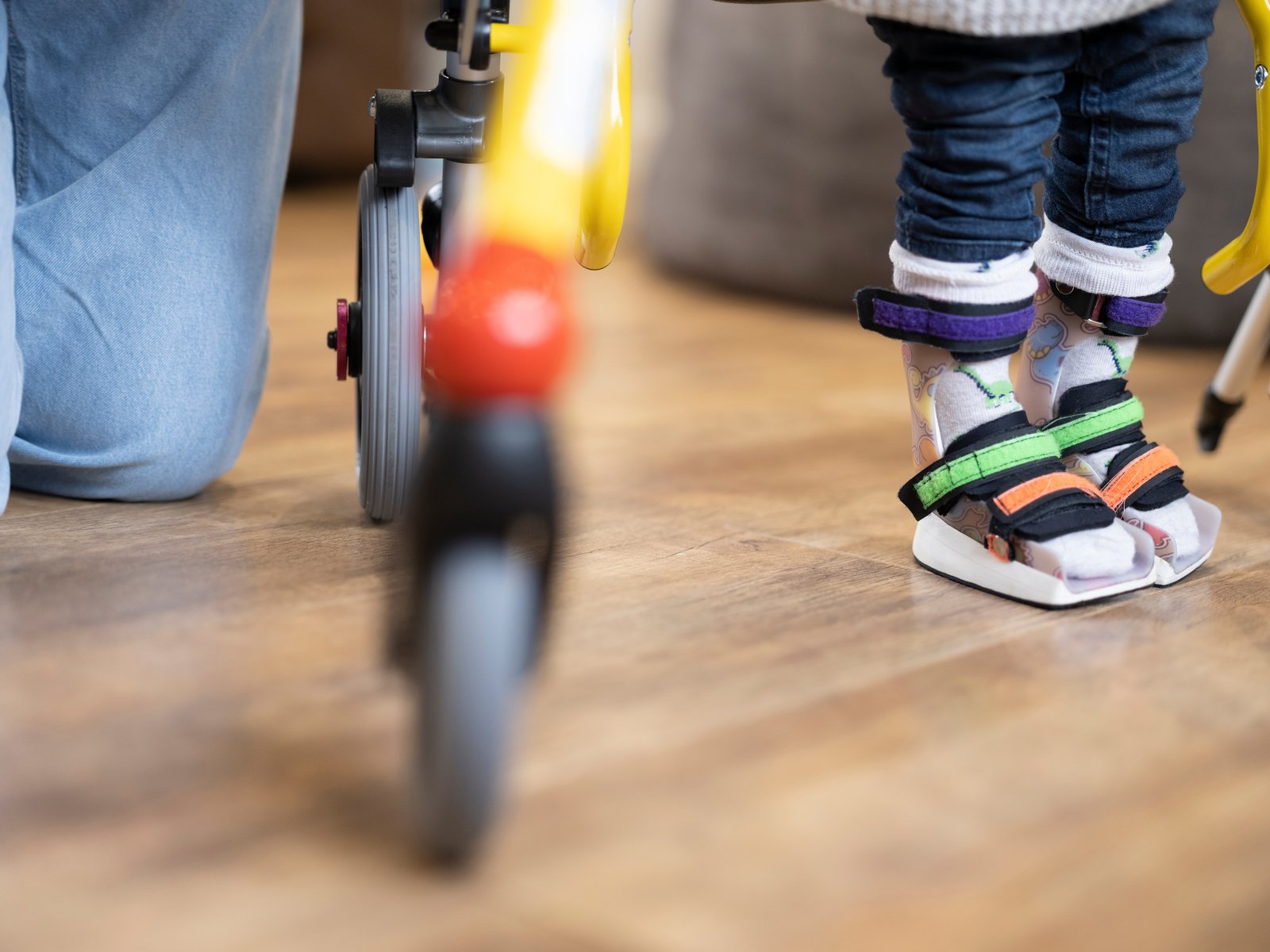 Kind mit Fußorthesen steht mit Hilfe eines Gehtrainers